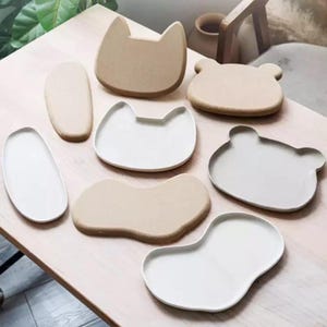 Peut inclure: Un ensemble de huit assiettes en céramique de formes et tailles variées, incluant des modèles en forme de chat, d'ours et des formes ovales irrégulières. Les assiettes présentent une finition mate lisse dans des tons neutres beige et crème.