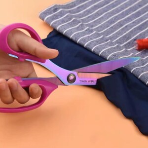 May include: Pink-handled scissors with iridescent blades cutting blue fabric. The scissors have the text "TWIN WOLF" printed on the blade. A striped fabric and a spool of red thread are also visible.