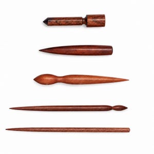 Puede incluir: Un conjunto de cinco herramientas de madera de color marrón oscuro con superficies lisas y pulidas. La colección incluye varias formas como una herramienta cilíndrica con extremo plano, una punta cónica, un centro bulboso, una aguja larga y fina, y una varilla recta.