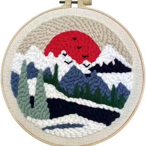 May include: A round, framed punch needle art piece featuring a mountain landscape with a red sun setting behind snow-capped mountains. The artwork includes blue, green, white, and black elements, with three birds flying across the sun.