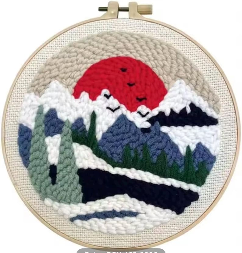 May include: A round, framed punch needle art piece featuring a mountain landscape with a red sun setting behind snow-capped mountains. The artwork includes blue, green, white, and black elements, with three birds flying across the sun.