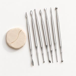 May include: A set of six double-ended stainless steel sculpting tools with various shaped tips, including ball, pointed, and flat ends, arranged vertically next to a single circular piece of light beige modeling clay with a curved indentation.