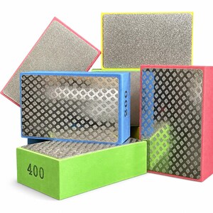 May include: A collection of rectangular diamond polishing blocks with foam bases in blue, green, yellow, and pink. The blocks feature metallic surfaces with diamond-patterned textures. The blue block is marked 200 and the green block is marked 400.
