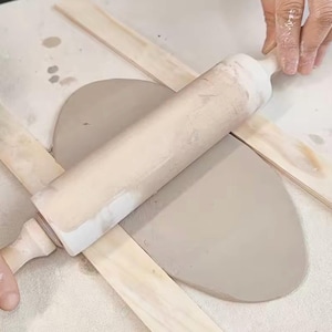 Puede incluir: Un rodillo de madera de color claro se utiliza para aplanar un trozo de arcilla gris. Dos tiras de madera se colocan a ambos lados de la arcilla para asegurar un grosor uniforme. La arcilla está sobre una superficie blanca. Se ven manos.