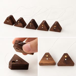 May include: Five dark brown wooden triangular tea bag holders with a smooth finish. Each piece features a small circular brass inlay and a thin silver-colored string attached to the top. The design includes a slot for securing tea bag strings.