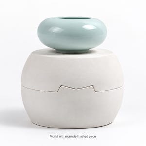 May include: A two-part white plaster casting mold with a rounded shape and a notched seam, topped with a smooth, glossy, light mint green ceramic bowl. The text "Mould with example finished piece" is printed at the bottom.
