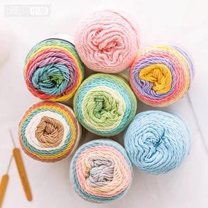 Könnte beinhalten: Eine Sammlung bunter Wollknäuel in Rosa-, Blau-, Grün-, Gelb- und Regenbogenfarben. Die Wolle ist kreisförmig angeordnet, mit zwei Häkelnadeln. Ideal für Strick- und Häkelprojekte.