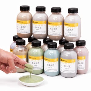 Puede incluir: Una colección de doce botellas de plástico transparente con tapas negras, cada una con 350 ml de barbotina líquida de colores. Las botellas tienen etiquetas amarillas con texto en chino. Un dedo sostiene una gota de barbotina verde sobre un cuenco.