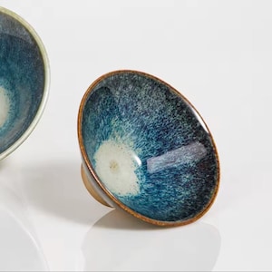 May include: Two ceramic bowls with a speckled blue and white interior and a brown rim. The bowls have a glossy finish and a small, rounded base. The bowls are set against a white background.
