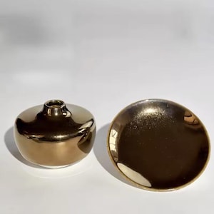 Könnte beinhalten: Eine goldfarbene Keramikvase und eine passende Schale. Die Vase ist rund mit einem schmalen Hals. Die Schale hat eine flache, kreisförmige Form. Beide Artikel haben eine glänzende, metallische Oberfläche.