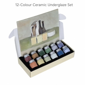 Puede incluir: Un set de 12 colores de bajo esmalte para cerámica en una caja rectangular con tapa abatible que muestra una pintura de bodegón. El conjunto contiene doce frascos de vidrio pequeños con tapas de rosca plateadas, cada uno con un color distinto.