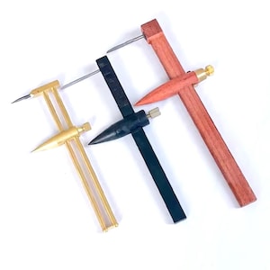 May include: Four different compass tools are displayed against a white background. One is gold-colored, one is black, and one is red. The tools have sharp points and are used for measuring and marking.