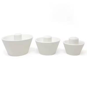 May include: A set of three white ceramic bowls, each featuring a central cylindrical pillar rising from the base. The bowls are arranged in descending order of size from left to right, all sharing a smooth, matte finish and a tapered, conical shape.