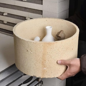 May include: A cylindrical, beige, porous ceramic kiln furniture ring with a rough, textured surface. Inside the ring, there is a smooth, white, narrow-necked ceramic vase and a small, brown, unglazed ceramic figurine with rounded features.