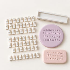 Pode incluir: Um conjunto de carimbos brancos individuais de letras e números, um cabo retangular branco, uma placa circular lavanda com letras maiúsculas e números em relevo, e uma placa retangular rosa claro com letras minúsculas em relevo.