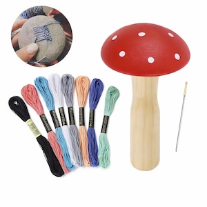 Puede incluir: Un cojín con forma de hongo rojo con lunares blancos, un tallo de madera y una aguja. También se muestra hilo de bordar de colores en varios tonos de rosa, negro, azul, blanco, gris, naranja y verde. Se muestra un pequeño cojín redondo y gris que se está utilizando.