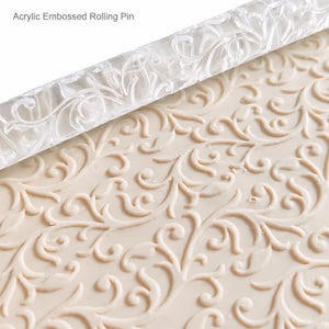 May include: A clear acrylic embossed rolling pin with a repeating floral vine pattern. The rolling pin is positioned above a beige surface that displays the same intricate, raised scrollwork design pressed into its smooth, flat texture.