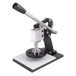 Puede incluir: Una máquina manual para hacer chapas con base de metal negro, plataforma rectangular blanca y mecanismo de prensa vertical plateado. El dispositivo incluye una palanca larga plateada con empuñadura negra, un troquel circular negro y pernos.