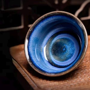 Könnte beinhalten: Eine handgefertigte Keramikschale mit einer auffälligen blauen Glasur. Das Innere weist ein wirbelndes Muster aus tiefen Blau- und Grüntönen auf, das einen einzigartigen visuellen Effekt erzeugt. Der Rand hat einen dunkelbraunen Rand, der Kontrast erzeugt. Die Schale steht auf einer Holzoberfläche.