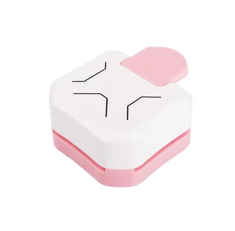 May include: A white and pink corner paper punch. The top is white with black corner guide lines. The bottom and the lever are pink. This craft tool is used for paper crafting and scrapbooking.