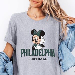 A woman wearing a grey t-shirt with a graphic of Minnie Mouse and the text "PHILADELPHIA FOOTBALL" printed on it.