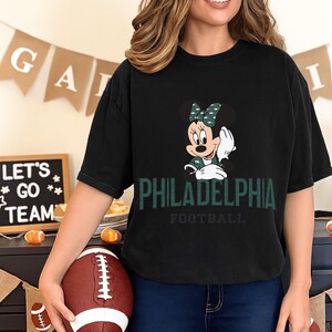 A woman wearing a black t-shirt with a Minnie Mouse graphic and the text "PHILADELPHIA FOOTBALL" is holding a football in front of a banner that says "LET'S GO TEAM".