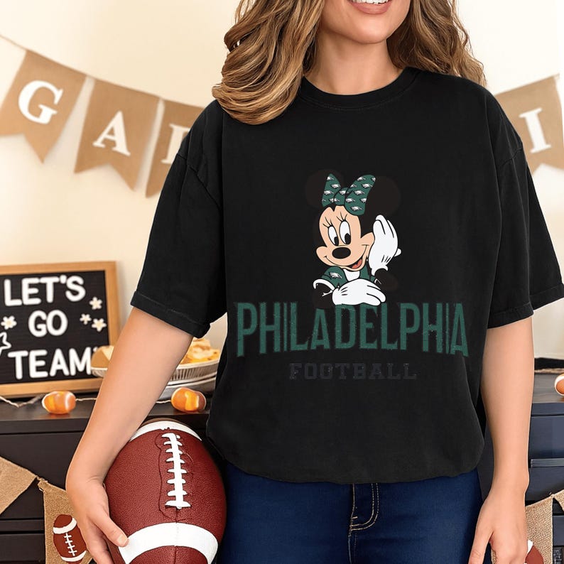 A woman wearing a black t-shirt with a Minnie Mouse graphic and the text "PHILADELPHIA FOOTBALL" is holding a football in front of a banner that says "LET'S GO TEAM".