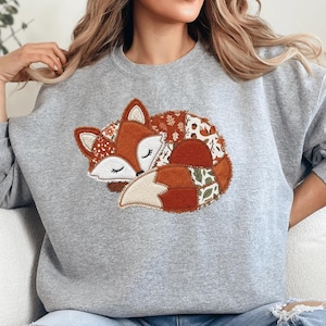 May include: A heather gray crewneck sweatshirt featuring a large, central applique of a sleeping fox. The fox is constructed from various fabric patches in shades of orange, cream, and brown, decorated with patterns of autumn leaves and acorns.