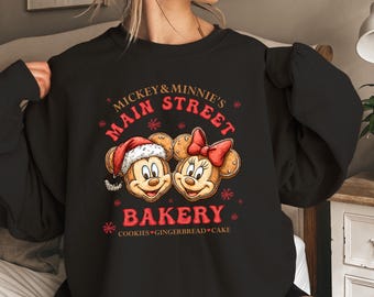 May include: Black sweatshirt featuring a festive design for Mickey & Minnie's Main Street Bakery. The graphic includes Mickey Mouse in a Santa hat and Minnie Mouse with a red bow, surrounded by snowflakes and the bakery's name. The text at the bottom reads "Cookies Gingerbread Cake."
