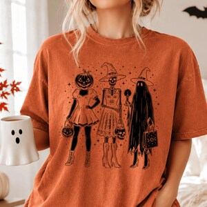 May include: A burnt orange short-sleeve t-shirt featuring a black graphic print of three figures: a pumpkin-headed person in a dress, a skeleton wearing a witch hat, and a ghost holding a candle and a bag, all surrounded by small stars.