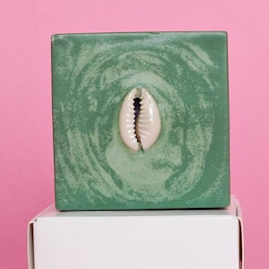May include: A square, green decorative object with a swirling pattern and a white seashell affixed to the centre. The object sits atop a white box against a pink background. The seashell is a focal point.