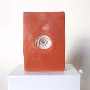 Può includere: Una scultura rettangolare color terracotta con un disegno a spirale bianco al centro. La scultura è esposta su una scatola bianca su sfondo bianco. L'opera d'arte ha una superficie strutturata.