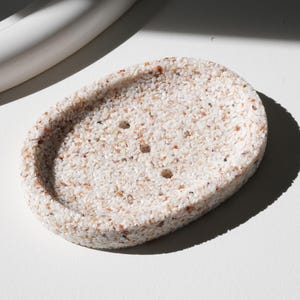 Può includere: Portasapone ovale con superficie maculata e strutturata. Il portasapone è principalmente bianco sporco con piccole macchie marroni e beige. Presenta tre fori di drenaggio e un bordo leggermente rialzato.