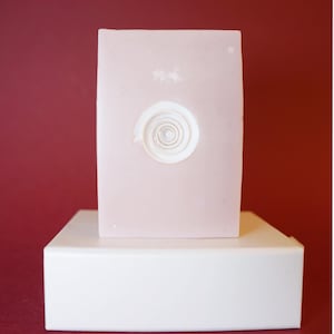 Könnte beinhalten: Eine hellrosa rechteckige Skulptur mit einem weißen Spiraldesign auf einem weißen rechteckigen Sockel. Der Hintergrund ist ein einfarbiger, kastanienbrauner Farbton.