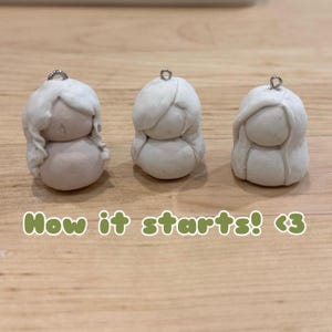 Puede incluir: Tres peque&ntilde;as figuras de arcilla blanquecina con lazos plateados. Cada figura tiene un cuerpo redondeado y una cabeza con pelo esculpido. El texto "How it starts! <3" est&aacute; en la parte inferior.