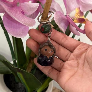 Puede incluir: Un llavero hecho a mano con una peque&ntilde;a figura de mu&ntilde;eca con cabello casta&ntilde;o y un vestido oscuro. El llavero tambi&eacute;n incluye una cuenta verde y un llavero plateado. Flores de orqu&iacute;deas rosas en el fondo.