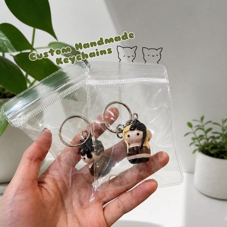 Puede incluir: Dos llaveros hechos a mano en bolsas de pl&aacute;stico transparente. Los llaveros presentan peque&ntilde;as figuras detalladas con cabello negro y rubio, y est&aacute;n unidos a llaveros plateados. El texto "Custom Handmade Keychains" es visible.