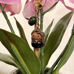 Puede incluir: Un llavero hecho a mano con una peque&ntilde;a figura de arcilla de una persona con cabello casta&ntilde;o y una parte superior negra. El llavero tambi&eacute;n incluye un dije verde. La figura est&aacute; unida a un llavero plateado.