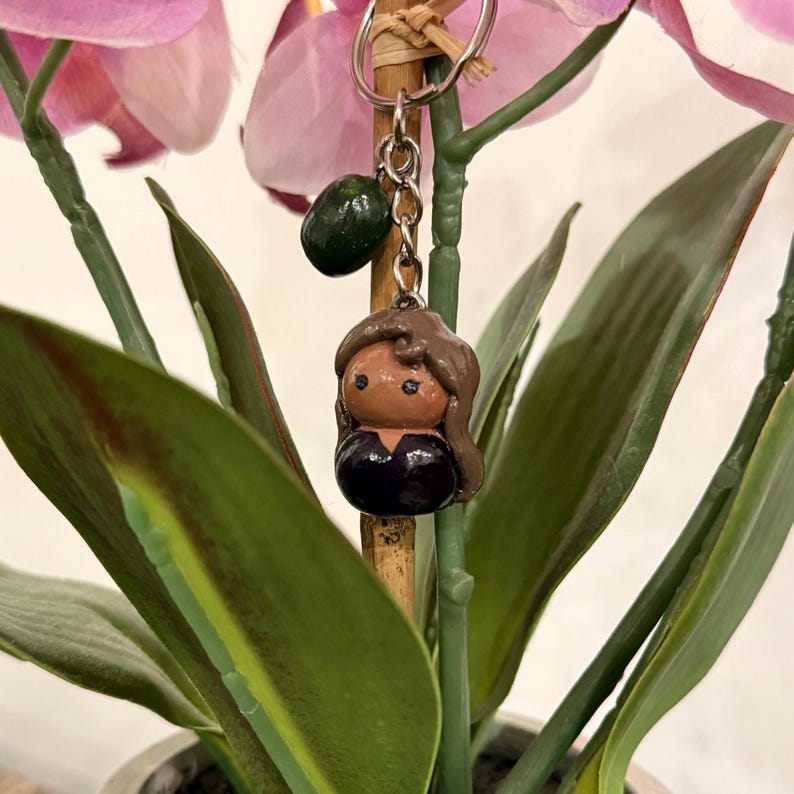 Puede incluir: Un llavero hecho a mano con una peque&ntilde;a figura de arcilla de una persona con cabello casta&ntilde;o y una parte superior negra. El llavero tambi&eacute;n incluye un dije verde. La figura est&aacute; unida a un llavero plateado.