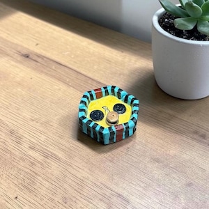 May include: A small, hexagonal ceramic trinket dish with a bright yellow interior and a rim featuring alternating vertical stripes of black, turquoise, and red. Two black buttons and one small tan button are embedded in the yellow base.