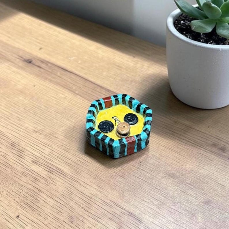 May include: A small, hexagonal ceramic trinket dish with a bright yellow interior and a rim featuring alternating vertical stripes of black, turquoise, and red. Two black buttons and one small tan button are embedded in the yellow base.