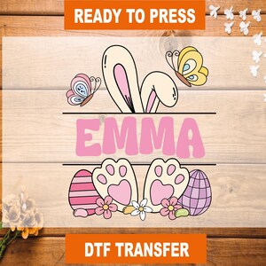 May include: A pink and white bunny ear design with the name "EMMA" in pink letters. The design includes butterflies, flowers, and Easter eggs. The text "READY TO PRESS" and "DTF TRANSFER" are also included.