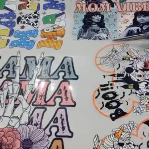 May include: Printed designs on a white surface, featuring the words "MOM VIBE" and "AMA". Other designs include a skeleton hand with flowers, a Halloween-themed design with a spider web, and various letter designs.