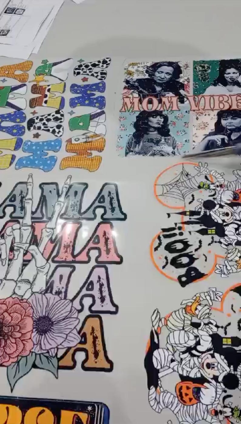May include: Printed designs on a white surface, featuring the words "MOM VIBE" and "AMA". Other designs include a skeleton hand with flowers, a Halloween-themed design with a spider web, and various letter designs.