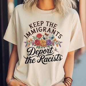 May include: A cream-coloured t-shirt featuring the text "Keep the Immigrants, Deport the Racists" in black script. The text is above and below a floral design with red, yellow, and purple flowers, butterflies, and a dragonfly.