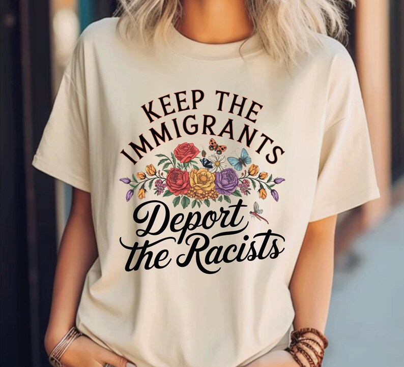 May include: A cream-coloured t-shirt featuring the text "Keep the Immigrants, Deport the Racists" in black script. The text is above and below a floral design with red, yellow, and purple flowers, butterflies, and a dragonfly.