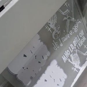 May include: A grey sheet with white graphics and text is inside a white machine. The sheet has a cloud-like shape with the text "If I was a bird" and other graphics including a skeleton and a bird.
