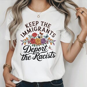 May include: White t-shirt featuring the text "Keep the Immigrants, Deport the Racists" in black script. The design includes a floral arrangement with red, yellow, and purple flowers, butterflies, and a dragonfly.