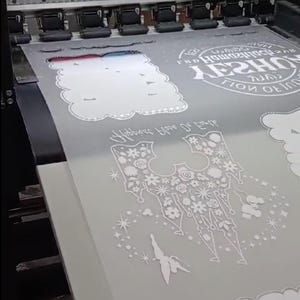 May include: A printer printing white designs on a grey sheet. The designs include floral patterns, cloud shapes, and text that reads "Happiness Blooms From Within". The printer is in operation, with the print heads moving across the material.