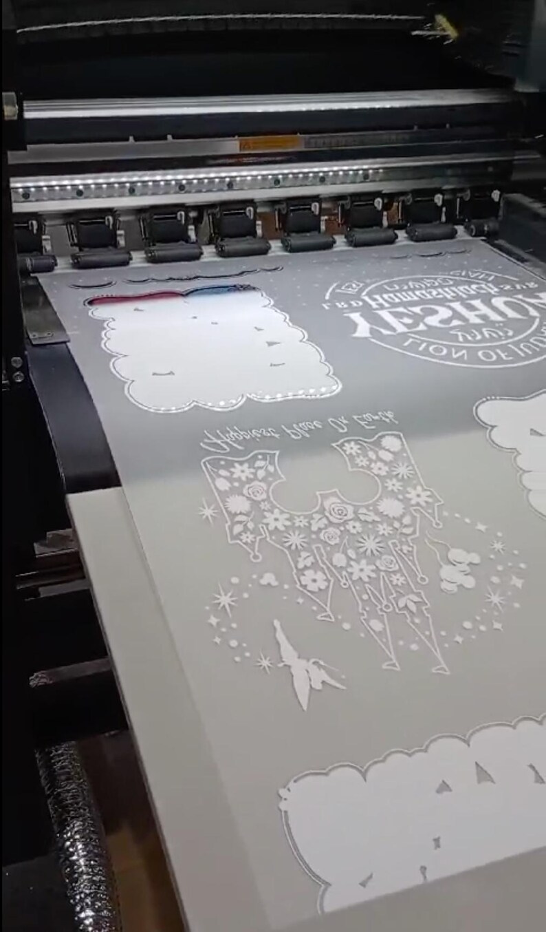May include: A printer printing white designs on a grey sheet. The designs include floral patterns, cloud shapes, and text that reads "Happiness Blooms From Within". The printer is in operation, with the print heads moving across the material.